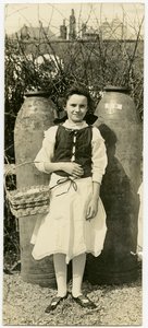 Lodret, sepia -fotografi, der viser en ung pige i en kjole, der står foran to store artilleri -skaller. Pigen holder en kurv i skækken i sin højre albue